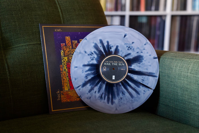 A marbled blue and black Hail The Sun splatter vinyl LP sits on a green chair, with its abstract city art album cover behind it. Blurred record shelves appear in the background. The LP releases October 2025.
