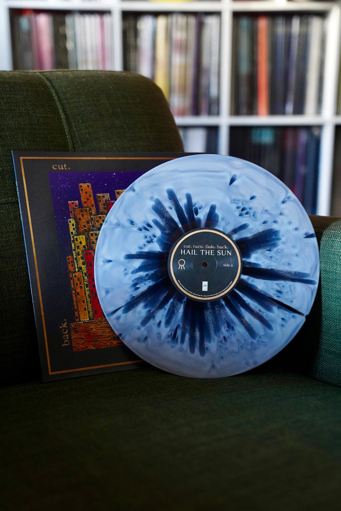 A blue and black splatter vinyl LP by Hail The Sun sits on a green chair before its vibrant album cover, with a blurred shelf of records behind. Album release October 2025.