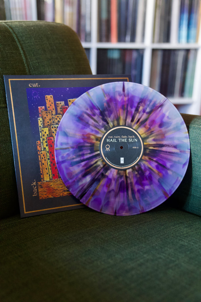 A purple & lavender starburst vinyl LP by Hail The Sun rests against its colorful album cover—abstract city buildings—on a green chair, with shelves of vinyl records behind. Album set for release in 2025.