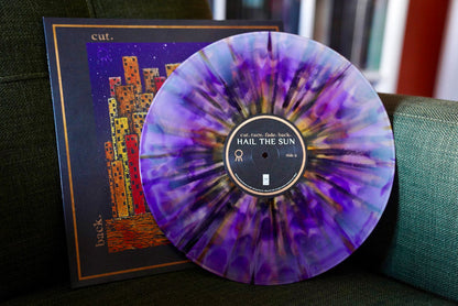 A marbled Purple & Lavender Starburst vinyl from Hail The Sun rests on a dark green chair, with its vibrant illustrated album cover and tall buildings visible in the background.