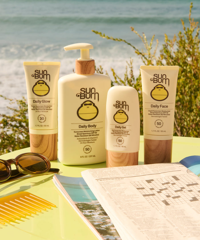 Sun Bum Daily 50 Face Lotion tube on a table with ocean background, sunglasses, and crossword puzzle nearby