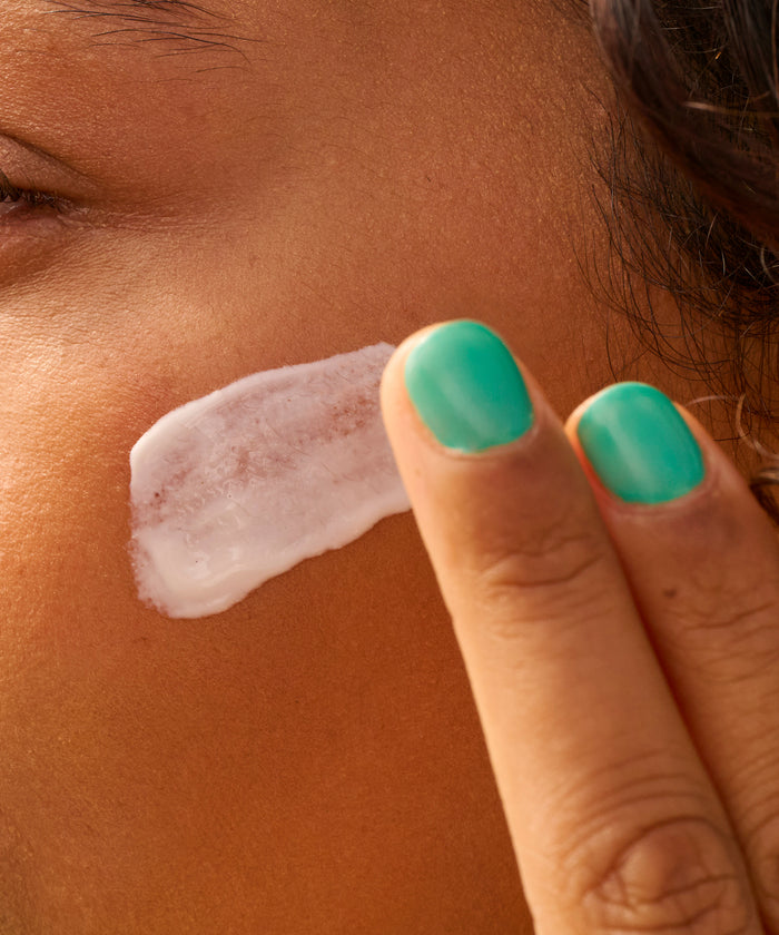Close-up of a person applying Daily 50 Face Lotion by Sun Bum on their cheek with teal nail polish on fingers