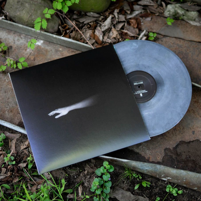 Image of Fairweather Deluge Vinyl LP with LP exposed to show color. Color of LP is Grey Smoke. Album cover shows half a ghostly arm on a black background. Both are laying on the ground on top of dirt and weeds.