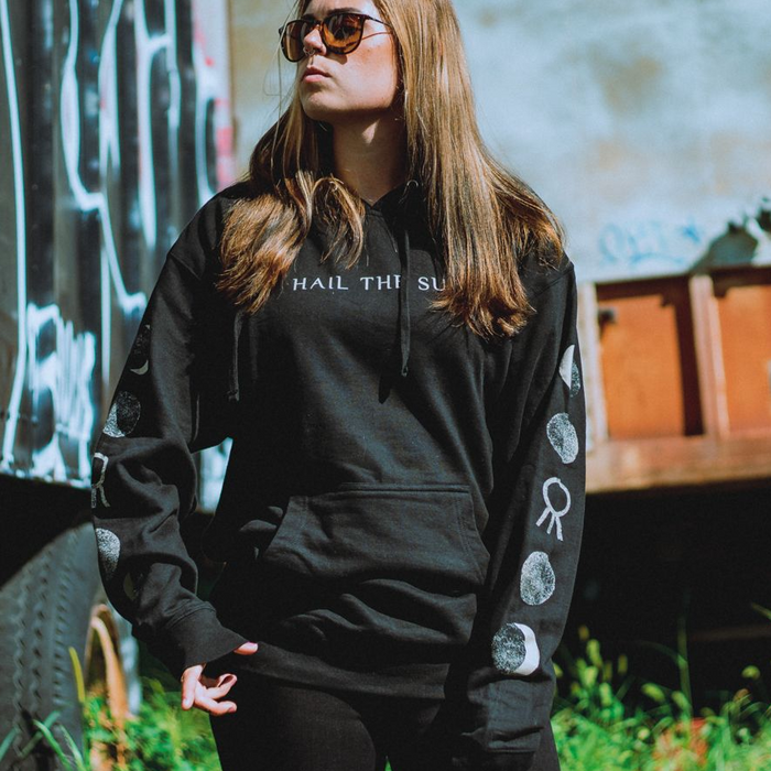 A woman with long, straight hair and sunglasses stands outdoors in a black Hail The Sun Divine Inner Tension Pullover Hoodie featuring moon phase sleeve designs, looking up and to the side.