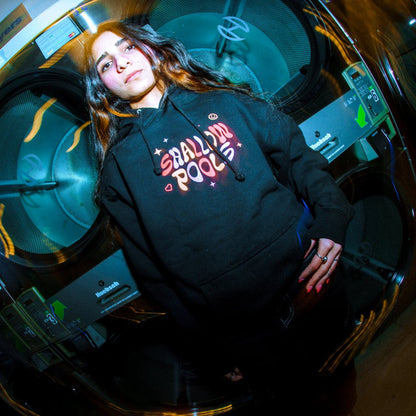 A woman with long hair stands by large washers, wearing a Shallow Pools pullover hoodie with "Small Pool" in pink and red. The photo uses a fisheye lens effect and vibrant, colorful lighting.