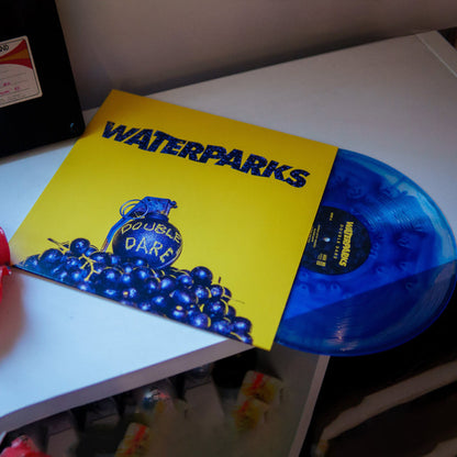 A Clear and Blue Cloud vinyl record partially slides out from a bright yellow Waterparks LP cover, which displays a hand grenade surrounded by blueberries with Double Dare written on the grenade.