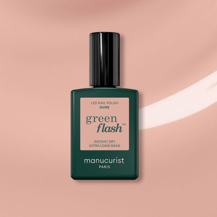 A bottle of Manucurist Dune Green Flash gel nail polish—a dark green shade with black cap and beige-pink label—stands against a soft pink background with a white streak, offering a modern twist on sandy beige nails.