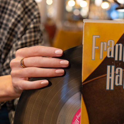 A hand with neatly manicured nails painted in Manucurist’s Dune shade holds a vinyl record beside a yellow album cover reading “Fran,” set against a softly blurred background with warm lights.
