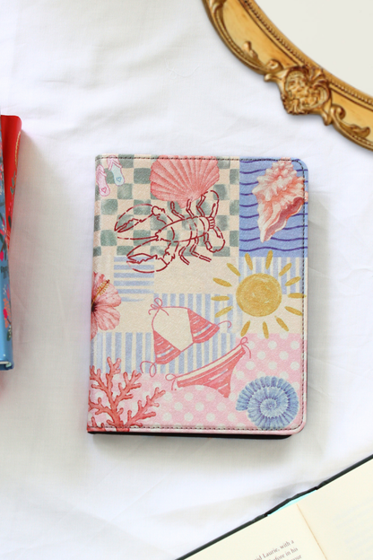 A kindle case featuring a colorful nautical patchwork print with a lobster, seashells, sailboat, sun, and coral motifs, displayed on a white fabric surface.