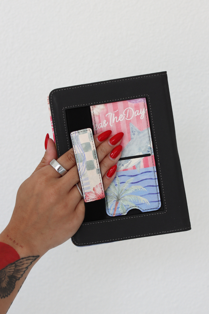 A hand with red nail polish holds a kindle case with colorful printed kickstand and hand strap, against a plain white wall