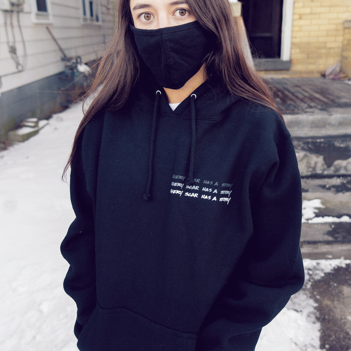 Black pullover hoodie with text that says EVERY SCAR HAS A STORY in white text in the top corner, written 3 times stacked on top of one another. An individual is modeling the hoodie and standing in front of two buildings, and there is snow on the ground.