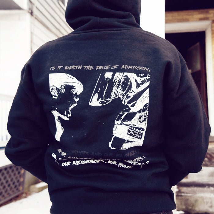 On the back, there is a drawing of a boy yelling in the face of a police officer that is much taller than him. Below that drawing is more text that is stacked in gradient coloring, getting more visible toward the bottom of the hoodie. An individual is modeling the hoodie, standing in front of two buildings. There is snow on the ground.
