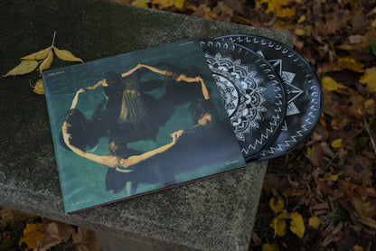 A Limited Edition Vinyl by Night Verses with a black and white geometric design partially slides out of its album cover, which shows women in black dresses holding hands in a circle, all resting on a stone bench amid fallen autumn leaves.