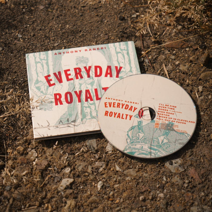A CD and its case from Anthony Raneri rest on dry, rocky ground amid brown grass. The case’s green and red artwork stands out, evoking the classic style of vintage album covers.