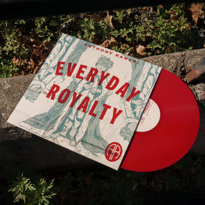 A red vinyl peeks out of an ivory jacket. There is a muted green print of a boy dressed up as a king with the words "Everyday Royalty" over him. The vinyl and jacket rests on a cement curb outside.