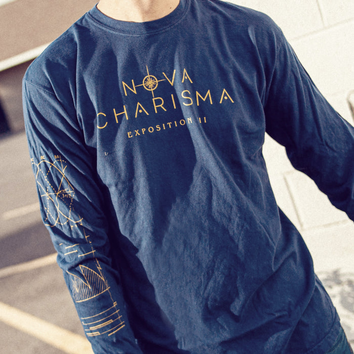 Navy long sleeve shirt with NOVA CHARISMA written across the chest. Inside the letter O of NOVA, there is a starlight design coming out of it. Below NOVA CHARISMA, there is smaller text that says EXPOSITION III. On one of the sleeves, there are various designs of graphs and data. An individual is modeling the shirt and standing in front of a building and in a parking lot.