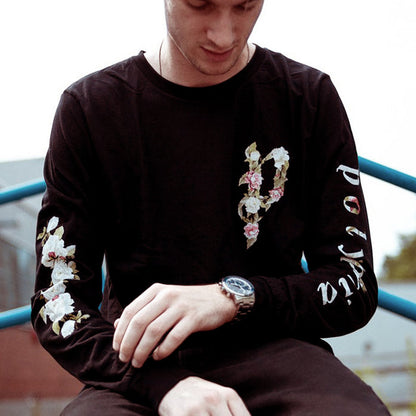 Black long sleeve shirt with the letter P written in the top right corner. The letter P is written using pink and white flowers, as well as some green leaves. The same design is on the back of the shirt, except much larger, covering the entire back. One sleeve says POLYPHIA on it, and the other has white flowers down the side. An individual is modeling the shirt, leaning against a blue railing.