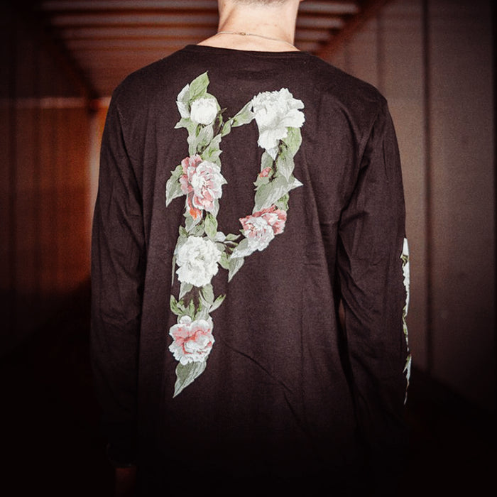 Black long sleeve shirt with the letter P written in the top right corner. The letter P is written using pink and white flowers, as well as some green leaves. The same design is on the back of the shirt, except much larger, covering the entire back. One sleeve says POLYPHIA on it, and the other has white flowers down the side. An individual is modeling the shirt, standing inside of a truck.