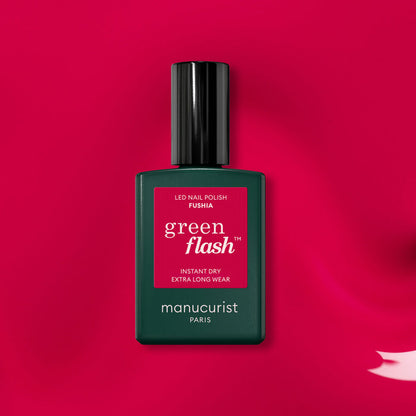 A bottle of Manucurist Fuchsia Green Flash gel polish rests on a vivid pink surface, with a swipe of fuchsia gel polish shown in the lower right corner.