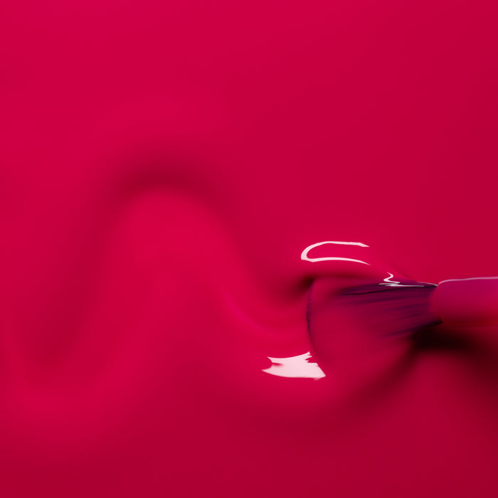A close-up of a brush spreading Manucurist Fuchsia gel polish in a wavy line on a smooth surface, with glossy reflections highlighting the vibrant pink color.