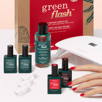 A hand with red nails rests beside Manucurist's Gift Set, featuring Green Flash gel polish, remover, a LED travel lamp, nail clips, and box, all arranged on a light surface.