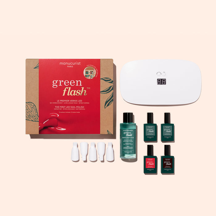 The Manucurist Gift Set includes a red and brown Green Flash box, a white LED travel lamp, four gel polishes in green and red shades, remover, and four white nail clips, all displayed on a light pink background.