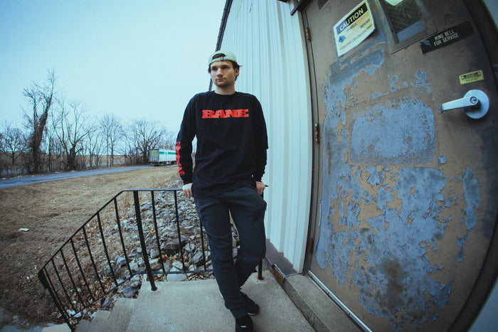 A young man in a black Bane sweatshirt, comfy printed apparel, dark pants, and a backwards cap stands on concrete steps near a worn metal door outside an industrial building on a cloudy day.