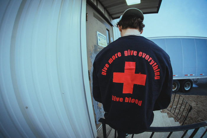 A person outside near a building and truck wears a dark Bane Comfort Colors shirt with a large red cross and “give more give everything give blood” printed on the back.