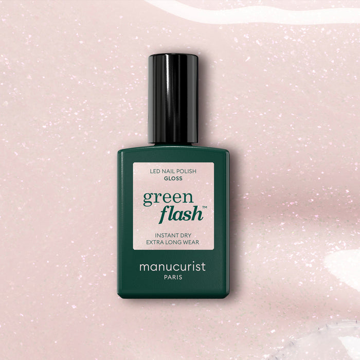 A dark green bottle of Manucurist Gloss gel polish rests on a pale pink, iridescent shimmer background. The label emphasizes instant dry, extra long wear, and high gloss.