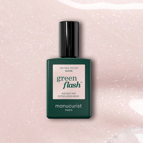A dark green bottle of Manucurist Gloss gel polish rests on a pale pink, iridescent shimmer background. The label emphasizes instant dry, extra long wear, and high gloss.