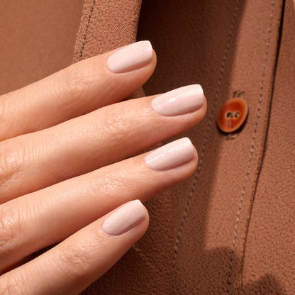 A close-up of a hand with neatly manicured nails painted in Manucurist Gloss, an iridescent pink polish, held against brown fabric with a visible button.