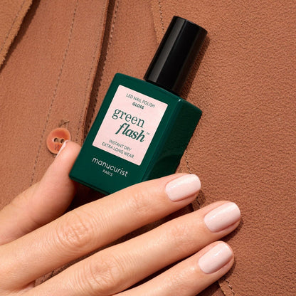 A hand with light pink, iridescent-shimmer polished nails holds a green bottle of Manucurist Gloss polish against a brown textured fabric background.