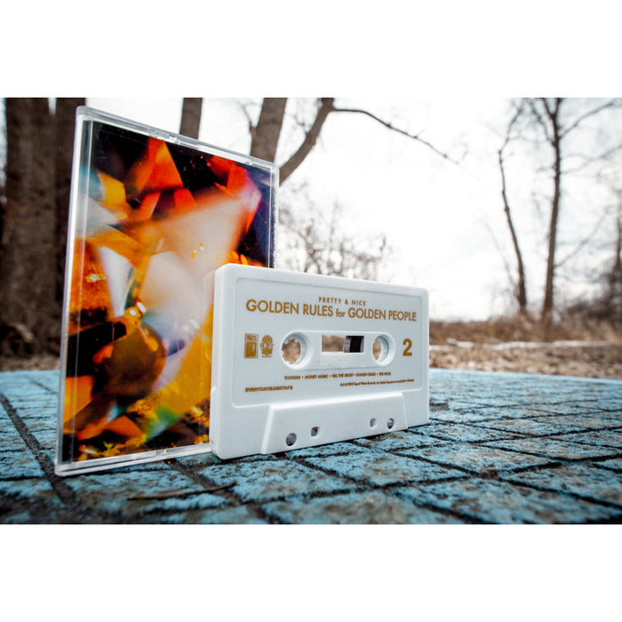 A white cassette tape labeled Golden Rules for Golden People by Pretty & Nice sits on a blue textured surface next to a colorful, abstract CD case with bare trees and a cloudy sky in the background, evoking nostalgia.