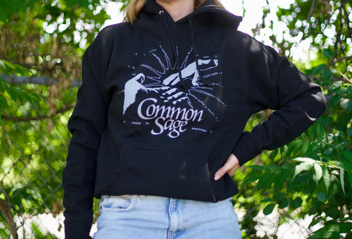 A person stands outdoors in a black Common Sage pullover hoodie paired with light blue jeans. Green leaves and sunlight are in the background.