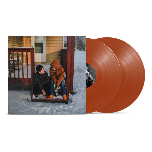 Two brown vinyl records from The Plot In You's "Happiness In Self Destruction" are placed next to an album cover featuring two people on a snowy porch and a terracotta-yellow house in the background.