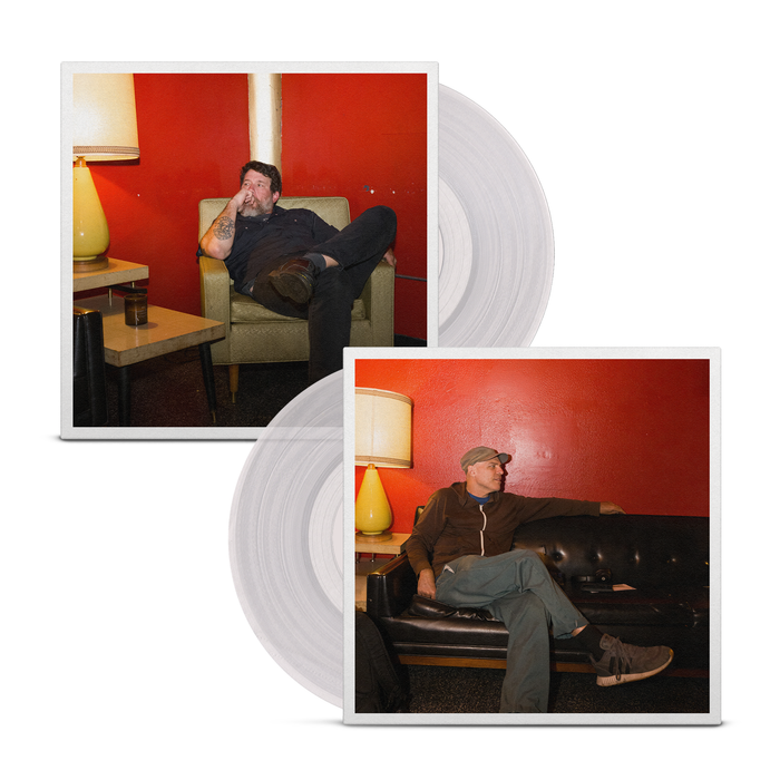A clear vinyl peeks out of a jacket that is dual sided. On one side a man is sitting on a yellow/cream chair behind a red wall and looking to the left. On the other side, a man in a baseball cap is sitting on a black couch in the same room and looking to the right.