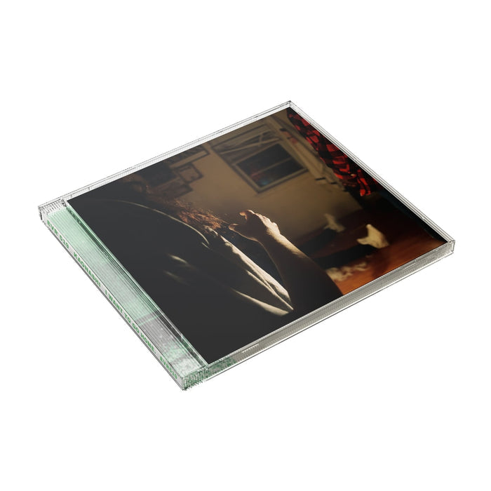 A CD showcases the image of three Kaonashi members in front of a window in a house with broken down blinds. The closest member is only showing his arms and hair, with the second only showing his red flannel, and the third sitting down with white shoes.