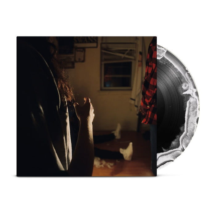 A black vinyl with a white crescent moon peeks out of a jacket with the image of three Kaonashi members in front of a window in a house with broken down blinds. The closest member is only showing his arms and hair, with the second only showing his red flannel, and the third sitting down with white shoes.