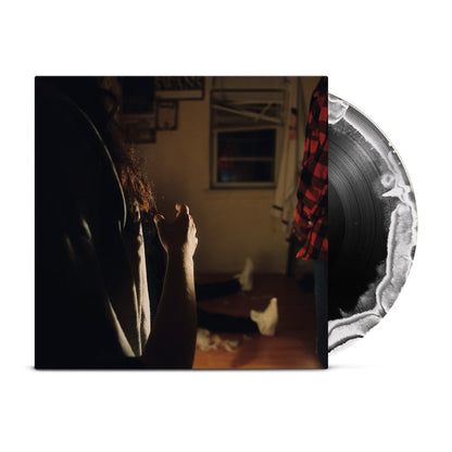 A black vinyl with a white crescent moon peeks out of a jacket with the image of three Kaonashi members in front of a window in a house with broken down blinds. The closest member is only showing his arms and hair, with the second only showing his red flannel, and the third sitting down with white shoes.