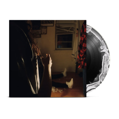 A black vinyl with a white crescent moon peeks out of a jacket with the image of three Kaonashi members in front of a window in a house with broken down blinds. The closest member is only showing his arms and hair, with the second only showing his red flannel, and the third sitting down with white shoes.