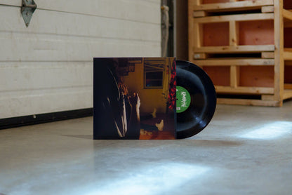 A black vinyl with white crescent moon peeks out of a jacket with the image of three Kaonashi members in front of a window in a house with broken down blinds. The closest member is only showing his arms and hair, with the second only showing his red flannel, and the third sitting down with white shoes.
