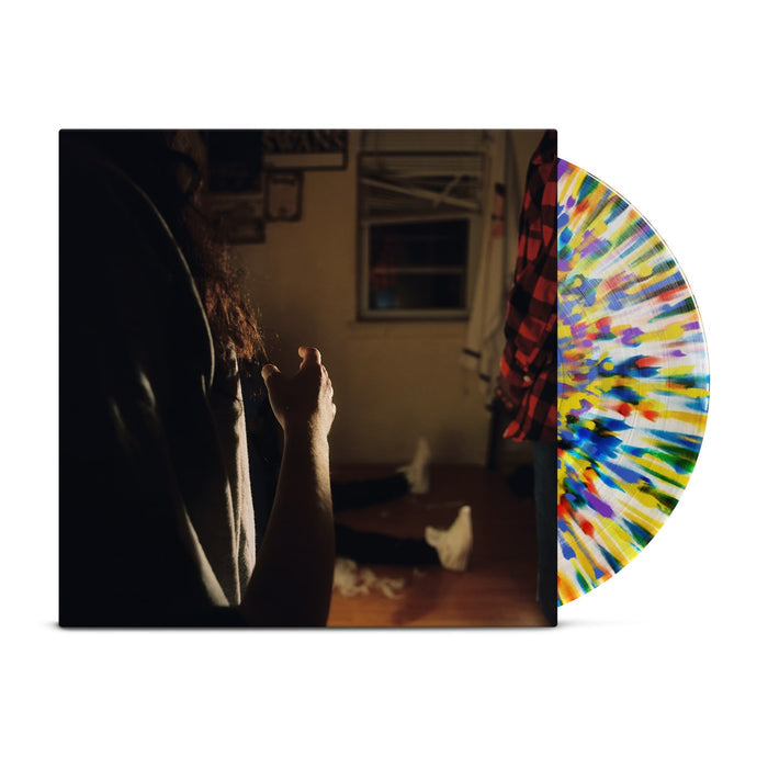 A rainbow splatter vinyl peeks out of a jacket with the image of three Kaonashi members in front of a window in a house with broken down blinds. The closest member is only showing his arms and hair, with the second only showing his red flannel, and the third sitting down with white shoes.