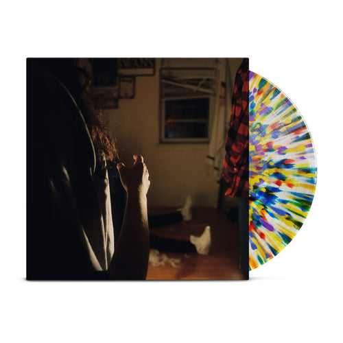 A rainbow splatter vinyl peeks out of a jacket with the image of three Kaonashi members in front of a window in a house with broken down blinds. The closest member is only showing his arms and hair, with the second only showing his red flannel, and the third sitting down with white shoes.