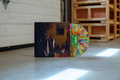A rainbow splatter vinyl peeks out of a jacket with the image of three Kaonashi members in front of a window in a house with broken down blinds. The closest member is only showing his arms and hair, with the second only showing his red flannel, and the third sitting down with white shoes. The vinyl is propped up in front of a garage door inside of a warehouse.