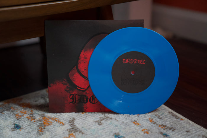 A vibrant blue vinyl record from Dwellings, featuring "Just A Man," partially covers its dark red-and-black album cover, both sitting on a patterned carpet with a wooden chair leg in the background.