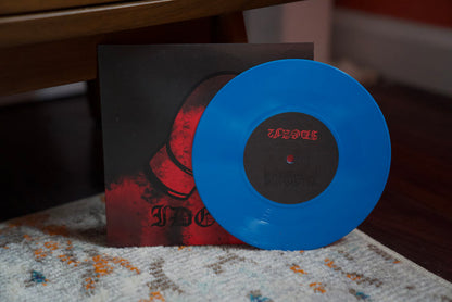 A vibrant blue vinyl record from Dwellings, featuring "Just A Man," partially covers its dark red-and-black album cover, both sitting on a patterned carpet with a wooden chair leg in the background.