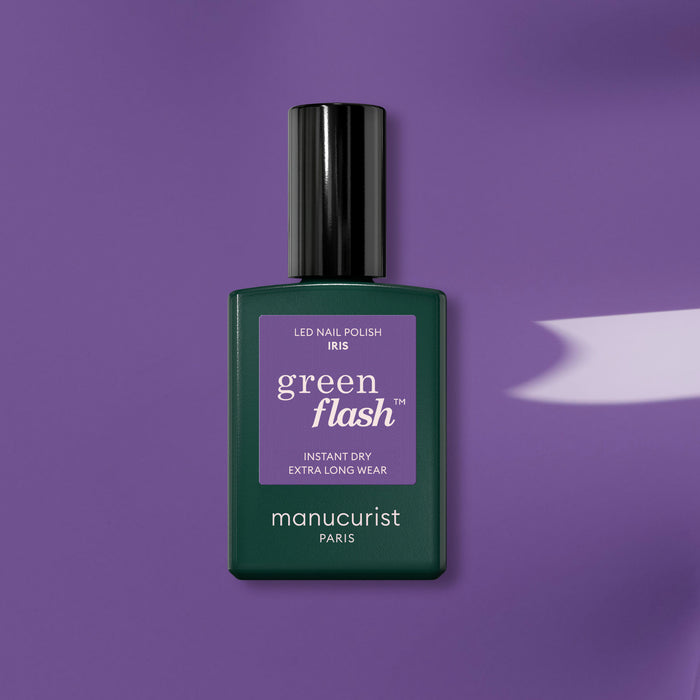 A bottle of Manucurist "Iris" gel polish rests on a purple surface, casting a gentle shadow. The dark green base and black cap complement the vibrant purple label, evoking blue undertones in this eye-catching Manucurist shade.