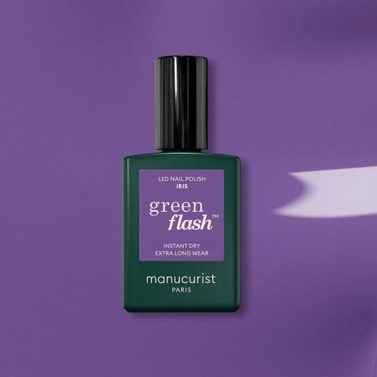 A bottle of Manucurist "Iris" gel polish rests on a purple surface, casting a gentle shadow. The dark green base and black cap complement the vibrant purple label, evoking blue undertones in this eye-catching Manucurist shade.