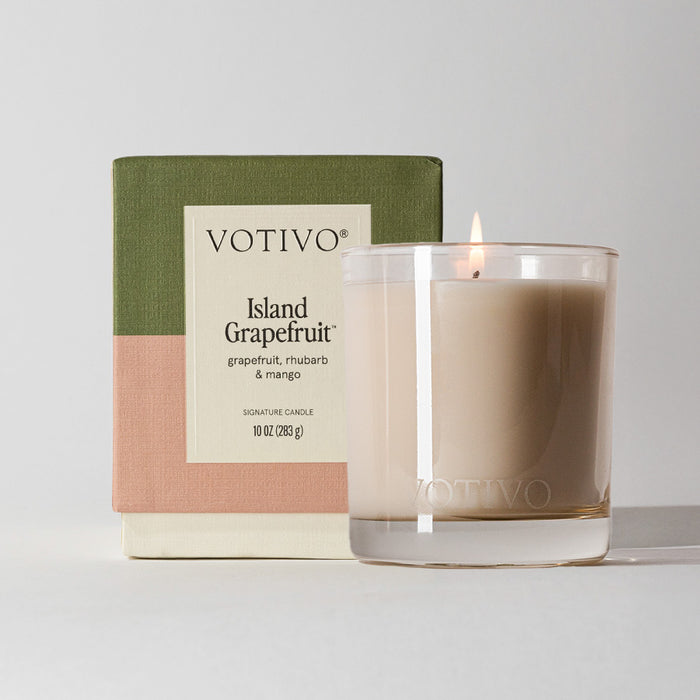 A lit Votivo Island Grapefruit 10 oz. Signature Candle, crafted from soy blend wax, sits in a clear glass holder in front of its green, pink, and beige color-blocked box featuring product details on the front.