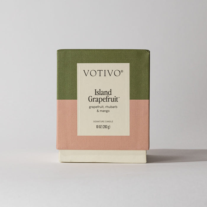 The Votivo Island Grapefruit 10 oz. Signature Candle, crafted with soy blend wax and featuring notes of grapefruit, rhubarb, and mango, comes boxed in green, pink, and cream color blocks against a light background.